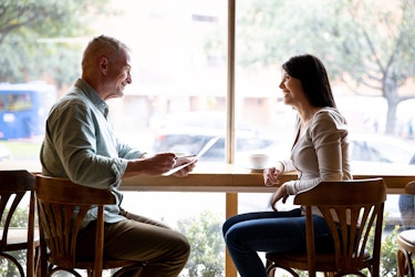  A cafe owner and a prospective employee sit at a table and two chairs in front of a window. The man smiles at the woman and holds her resume in his left hand. 