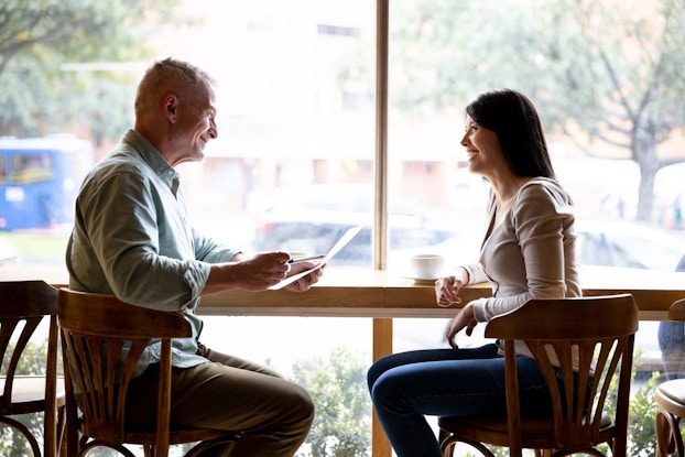  A cafe owner and a prospective employee sit at a table and two chairs in front of a window. The man smiles at the woman and holds her resume in his left hand.