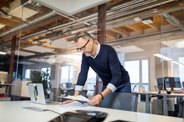  A wide shot of a man in glasses leaning over a conference room table to study some papers, one of which he holds in his left hand and the rest spread out on the table next to an open laptop. The man has a gray beard and dark blond hair graying at the temples. He wears jeans and a navy sweater over a white collared shirt. The conference room walls behind the man are made of transparent glass, and through them can be seen a warehouse space converted for office use, with desks arranged in pairs facing each other.