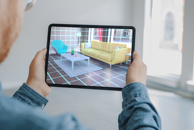  An over-the-shoulder shot of a man looking at a room through an electronic tablet. The room in the background is empty, but the room on the tablet screen is covered in a grid, with a couch, rug, coffee table, chair, lamp and wall art projected via augmented reality.