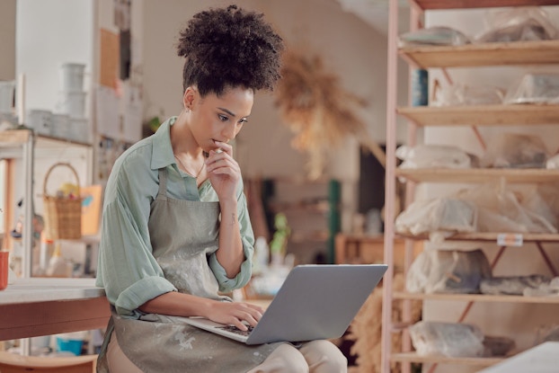  Woman artist who is thinking and working on a laptop in her studio.