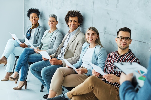  A line of five people of varying ethnicities and genders sits in a line of chairs against a light blue marble wall. All five people look expectantly and smiling at the person in the foreground, who is out of focus and partially off-screen. The only part of this person that can be seen is one arm, which holds a clipboard.