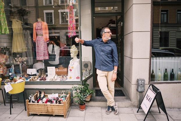 A vintage clothing store owner stands in front of his store, looking away from the camera, with a variety of items laid out on the sidewalk in front of his store for customers to peruse.