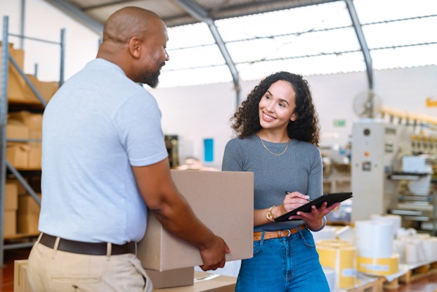 A woman writes something on a clipboard as she smiles at a man standing next to her with a cardboard box in his hands. The woman has shoulder-length curly hair and is wearing a gray shirt and jeans. The man is bald with a goatee and he wears a light blue shirt and khakis. In the background is a large warehouse space with shelves of cardboard boxes.
