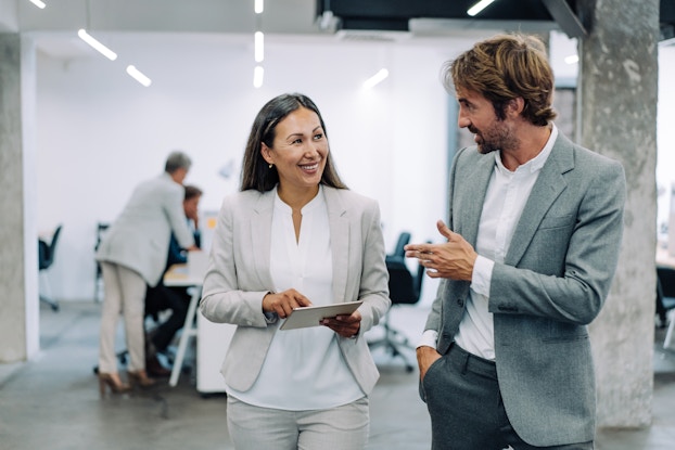 A man and a woman, both wearing light gray suits over white shirts, are standing next to each other and conversing. The man is speaking while gesturing and the woman is holding an electronic tablet. Behind them, in a large room lit by rows of overhead lights, is a set of connected office desks and several wheeled chairs. Two people appear to be huddled around one of the desks.