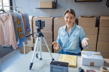  A woman sits at a table in a storage room in front of rows of stacked cardboard boxes. She speaks to a smartphone mounted on a tripod and gestures at the mailing paraphernalia (manila envelopes, boxes, tape, a stamp printer) on the table in front of her. 