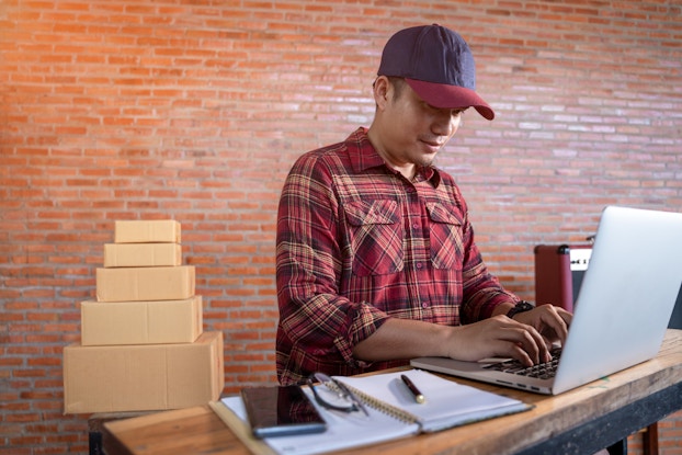  A young man in a baseball cap and a red plaid shirt stands at a wood-topped counter and types on a laptop. Behind the man is a brick wall and a stack of cardboard boxes.