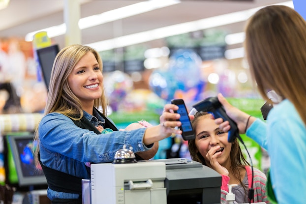  A smiling woman in a chambray shirt holds up a smartphone in front of a cashier, who faces away from the camera and scans the phone screen with a handheld scanner. A young girl with braces is standing next to the customer and smiling with one hand held near her mouth. The customer also has a baby in a carrier stapped to her chest.