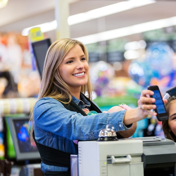 A smiling woman in a chambray shirt holds up a smartphone in front of a cashier, who faces away from the camera and scans the phone screen with a handheld scanner. A young girl with braces is standing next to the customer and smiling with one hand held near her mouth. The customer also has a baby in a carrier stapped to her chest.