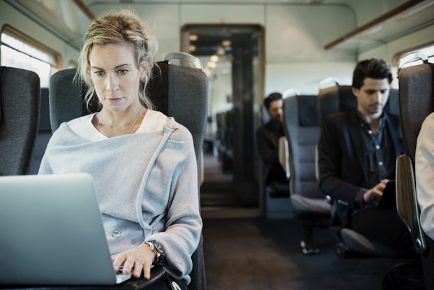 Several people sit in plush chairs inside of a train. Closest to the viewer, sitting in a seat on the left side of the train, is a woman with tied-back blonde hair. She wears an assymetrical pale blue blouse and looks at the open laptop in front of her with a look of concentration. One row back and sitting on the right side of the train is a man in a dark jacket. He looks down at something in his hands.