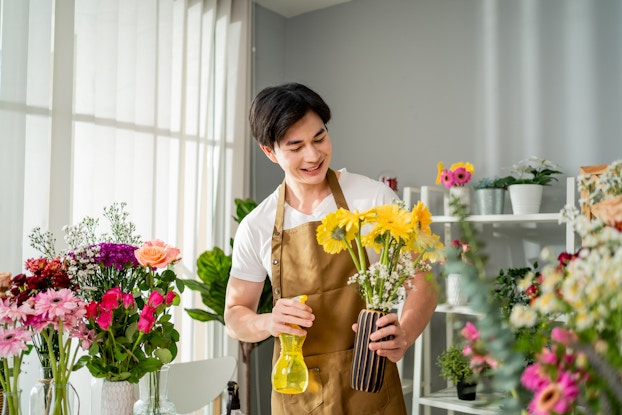 A young man in a flower shop holds a vase of flowers in one hand and sprays the yellow flowers with water from a yellow spray bottle. The man wears a white T-shirt under a brown apron.
