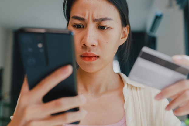 A young woman is holding her phone and looking at the screen with a concerned look on her face. She is holding a debit card in her left hand. She is reviewing a suspicious transaction in her account.
