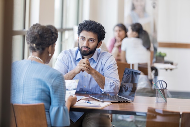 A man in a button-down shirt sits at a desk across from a woman reading from a pamphlet. A laptop is open on the desk beside them. In the background, two women sit across from each other at a similar desk.