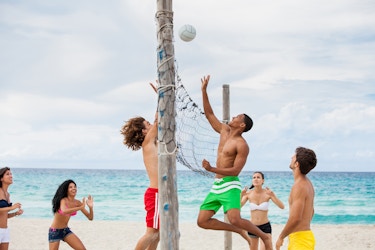  A group of young people plays volleyball at the beach. Two of the players, a long-haired man in red swim trunks and a short-haired man in light green swim trunks, are jumping up to the net, their hands outstretched to hit the mid-air volleyball. The four other players (three women in bikini tops and shorts and one man in yellow swim trunks) look on. In the background are the turquoise waters of the ocean and a pale, cloudy sky. 