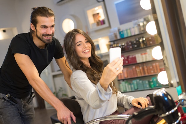  A woman uses her smartphone to take a selfie of herself and her hair stylist. The woman sits in the styling chair and has slightly wavy brown hair. The stylist behind her wears a black t-shirt and has hair clips clipped to the pockets of his jeans. He has a beard and a small man bun. In the background are several shelves of hair products sorted by color.