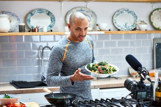  A man stands at a kitchen counter holding a bowl of mixed vegetables and speaking to the camera mounted in the foreground. The camera is a photographer's camera with a long microphone mounted on top of it. The man has a shaved head and stubble, and he wears a long-sleeved gray shirt with orange stripes around the shoulders. In the background is another kitchen counter with a sink. A shelf above this counter holds several porcelain plates.