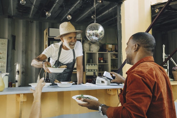 A man leans out of the front of a food stand to hand a bowl of food to a customer while speaking to another customer. The food stand worker wears a denim apron and a straw hat; he is older with a white beard. Behind him in the food stand are a glass-fronted refrigerator and shelves holding various cans; a disco ball and a string of small round lightbulbs hang from the ceiling. Outside of the food stand, a hand reaches from off-screen to take the offered bowl of food. A man in an orange shirt faces away from the camera and talks to the food stand owner. He is holding a bowl of food with one hand and holding his smartphone up to a digital point-of-sale device with the other hand.