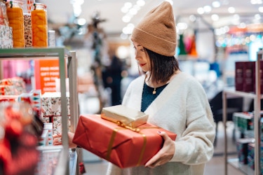  A young woman browses a shelf in a brightly lit store. The woman has shoulder-length dark hair and wears a tan hat and a pale blue sweater. She holds two wrapped packages, one wrapped in red paper and one in gold paper. 