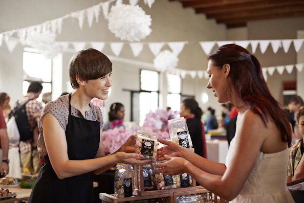 Two women stand in front of a display of bagged candies. The woman on the left, who has short brown hair and wears a black apron, hands a small bag of colorful teardrop-shaped candies to the long-haired woman on the right. In the background is a crowded shop with white bunting hanging from the ceiling.
