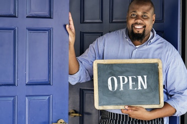  A Black man stands in front of a set of large blue doors. He smiles as he holds a sign that reads "OPEN." 