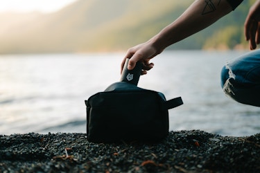  Image of a hand pulling a Black Wolf Nation product out of a toiletry bag on a lakeside. 