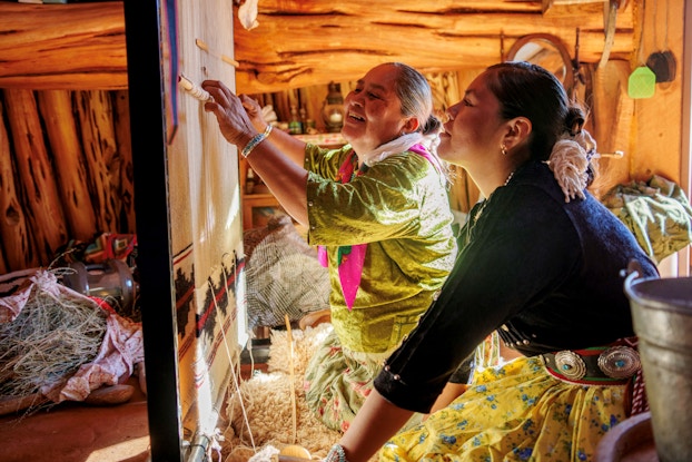 A Navajor grandmother sits before a loom with her grandaughter. They are weaving a blanket on a loom. The grandmother is showing her grandaughter how to make a Navajo blanket.