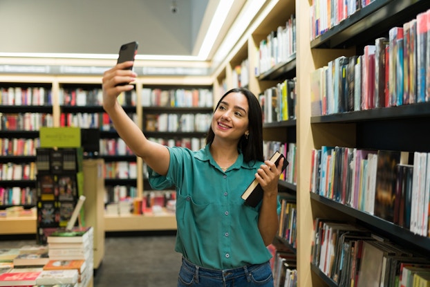 Woman smiling at her phone and taking a photo of herself holding a book inside of a bookstore.