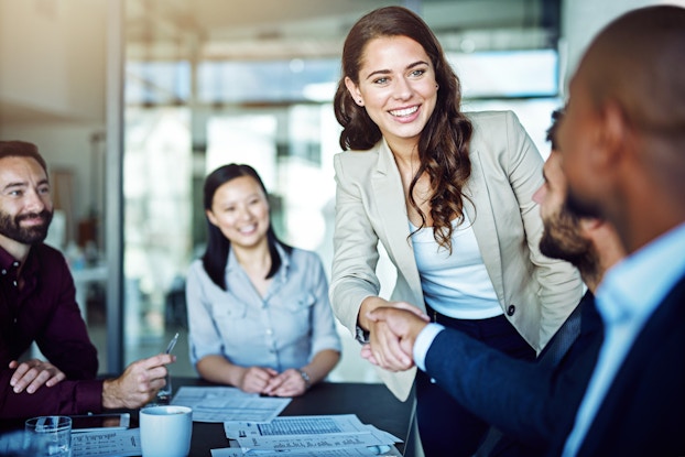 A group of people in business dress sit around a table in a glass-walled conference room. A woman standing up shakes the hand of one of the men at the table.