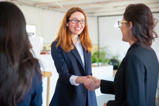  A red-haired woman in businesswear stands in an open office space and shakes hair with another woman dressed similarly. A third woman watches them, her back to the viewer.