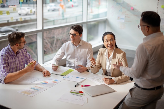 Four people sit around a square table and talk with each other. The two men on the left both wear glasses and have dark hair; they are in conversation with each other. On the right, a seated woman in a tan jacket speaks to a standing man who faces away from the viewer. The table is scattered with dry-erase markers and papers showing charts in shades of blue and orange.
