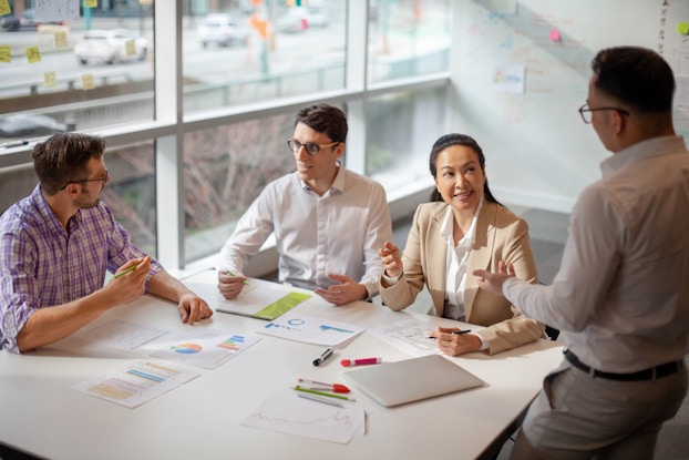  Four people sit around a square table and talk with each other. The two men on the left both wear glasses and have dark hair; they are in conversation with each other. On the right, a seated woman in a tan jacket speaks to a standing man who faces away from the viewer. The table is scattered with dry-erase markers and papers showing charts in shades of blue and orange.