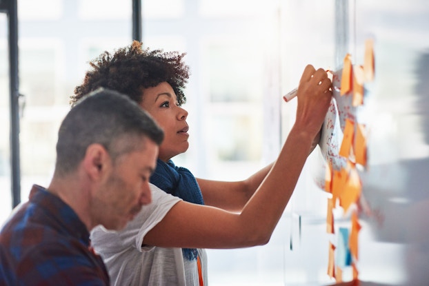 A businessman and businesswoman brainstorm on a whiteboard in a tech startup office.