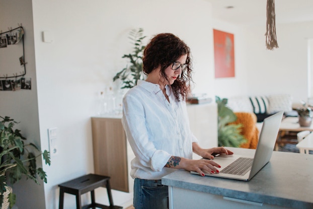 A young woman is pictured in her home working on a laptop computer that is positioned on a kitchen counter.