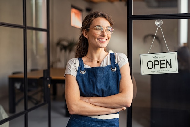 A young woman leans against the frame of a doorway. The door and the wall next to it are both made of glass with black windowpanes cutting the glass into rectangular sections. The glass wall against which the woman leans has a suction cup stuck to it. An open sign dangles from the suction cup. Behind the woman is a large room with cream walls. A table and chairs and a couple of tall plants stand inside.