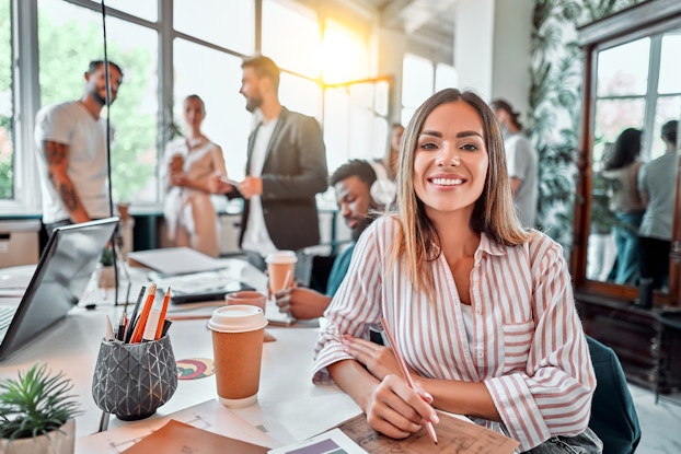  Smiling woman sitting at a desk inside an open-concept office with coworkers in the background.