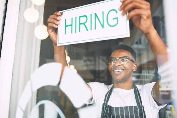A close-up shot of a smiling man in a striped apron, seen through the glass of a shop's window or door. The man's arms are raised as he attached a sign to the inside of the glass; the sign is white with the word "HIRING" in green.