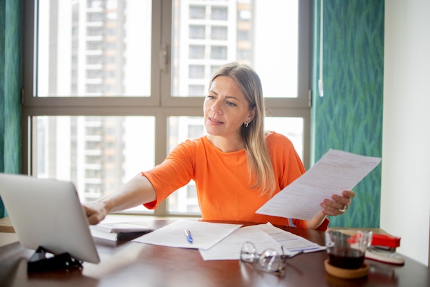 A woman in an orange shirt sits at a desk with a window at her back. She uses one hand to tap on the screen of an electronic tablet and holds a piece of paper in her other hand. The desk in front of her is scattered with more papers.