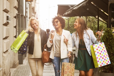 Group of three smiling friends walking outside and holding shopping bags. 