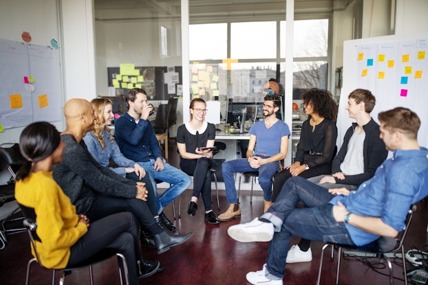 Nine smiling people of various ethnicities sit in a circle of chairs in an office meeting space. Behind them is a large set of floor-to-ceiling windows showing another office room. On either side of the windows is a whiteboard covered with sticky notes.