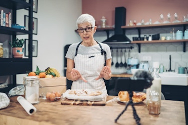  An older woman in an apron stands in a kitchen, explaining something as she's being filmed by a smartphone on a tripod. The woman is behind a large wooden counter with some flour-dusted dough on a tray directly in front of her. A crate of fruit, a container of sugar, and a trio of eggs sit to one side of the table, while a glass jug of milk and a plate of pastries sit off to the other side. The woman has short white hair and wears glasses, a white checkered apron, and a pale gray T-shirt. 