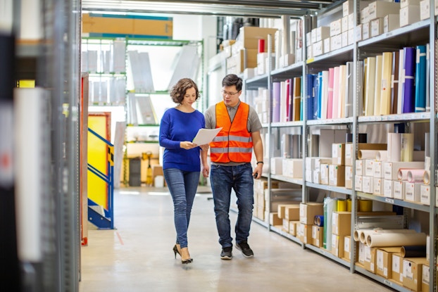 Two employees are walking between rows of shelving units in a warehouse. The person on the left, a woman with bobbed brown hair, is holding a packet of papers. The person on the right, a man wearing glasses and an orange high-visibility vest, looks over at the papers in his coworker's hands. The shelves in the background are filled with cardboard boxes, cardboard tubes, and multicolored folders.