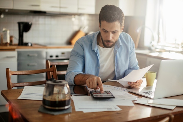  A bearded man sits at a kitchen table scattered with papers, an open laptop, and a half-full coffee carafe. With one hand, the man presses a key on a calculator. He holds a piece of paper in his other hand.