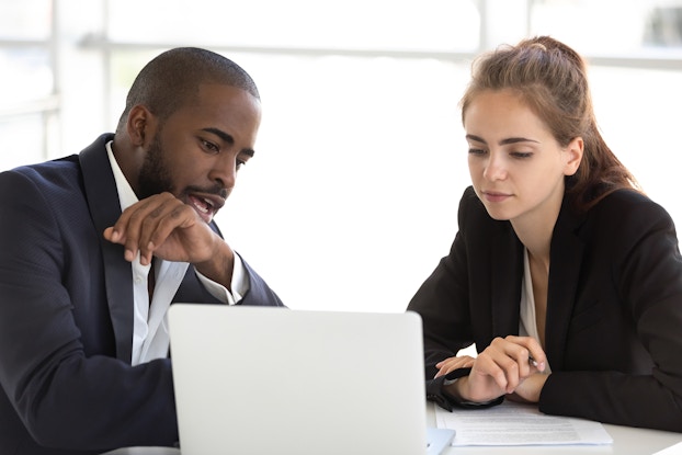  A man and woman, both wearing white shirts and black blazers, sit side by side at a table and look down at an open laptop. The man has a beard and shaved head, and his mouth is open as he speaks to the woman. The woman has dark blonde hair in a ponytail and holds a pen in one hand. In front of her on the table is a small pile of papers covered with text.