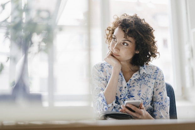 A woman sits in front of a large window and looks off to one side with a thoughtful expression. The woman has curly, shoulder-length hair and wears a white button-up shirt patterned with blue flowers. She holds a smartphone in one hand and has the other hand balled into a fist and held to her chin.