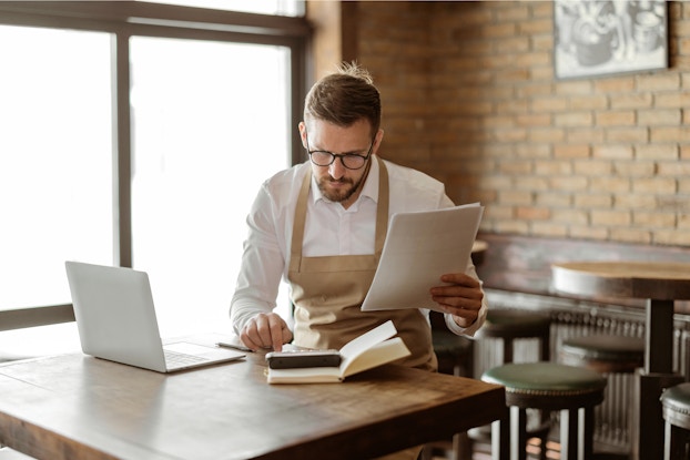  A bearded man in a tan apron sits at a table in a restaurant or cafe and types something on a calculator. The man holds a stack of papers in one hand and is looking down at the calculator with concentration. A laptop sits open on the table next to the calculator, which sits on top of an open book.