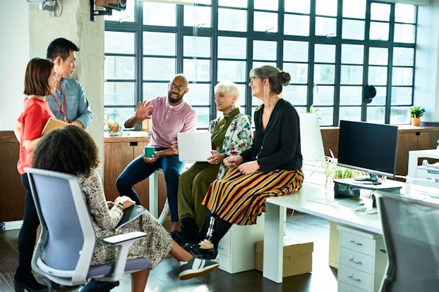  A group of six people of diverse ages, ethnicities, and genders sits and stands in a loose circle in an office setting. Everyone's attention is on the fourth person from the left, a bearded Black man, who is speaking and gesturing with one hand while holding a coffee mug with his other hand. He is sitting on top of a desk, as are an older woman holding a laptop and a woman with a prosthetic leg. Two other people are standing and one woman is sitting in a wheeled office chair.