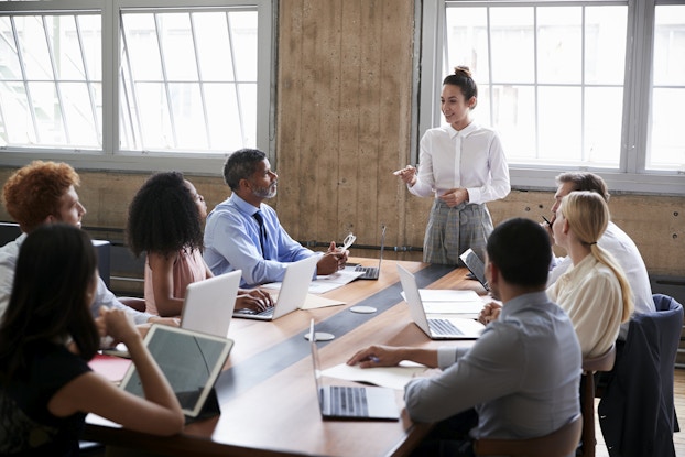 A group of people of varying ages, races and genders sit around a wooden conference table in a wood-paneled room. The seven people at the table watch a woman standing at the head of the table. The woman is wearing a white button-up shirt and gray plaid pants. She is speaking with a smile and pointing a finger in emphasis.