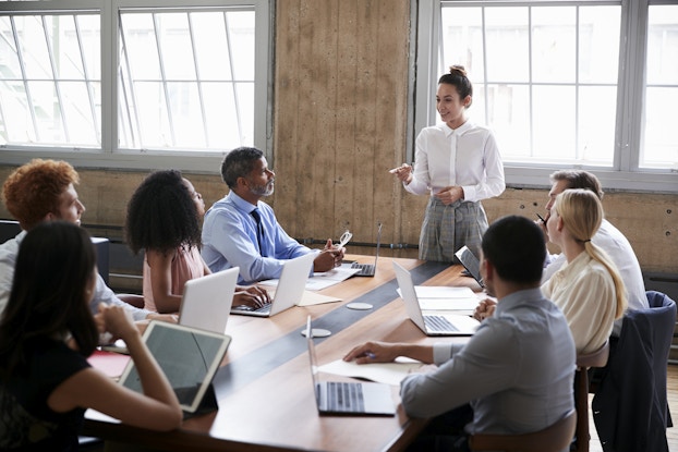  A group of people of varying ages, races and genders sit around a wooden conference table in a wood-paneled room. The seven people at the table watch a woman standing at the head of the table. The woman is wearing a white button-up shirt and gray plaid pants. She is speaking with a smile and pointing a finger in emphasis.