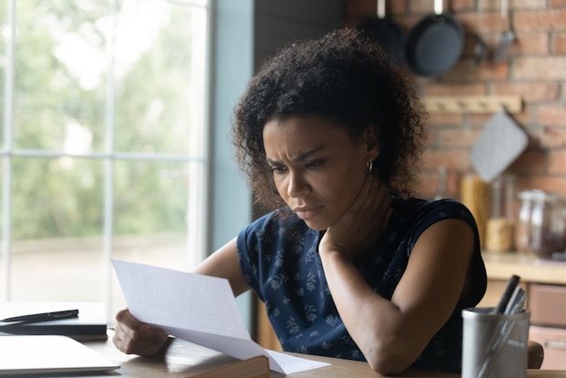 A woman sits at a table and looks at a piece of paper with a worried look. She has one hand to the side of her neck.