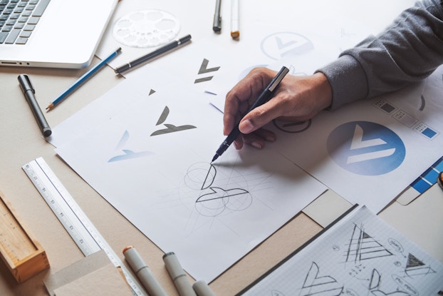 A piece of paper shows three versions of an A-shaped logo: a solid black version, a faded pale blue version and a sketched outline. A hand reaches in from out of frame holding a pen to add to the sketched outline. Scattered around the paper are pens, markers, a ruler and other papers bearing sketches of similar A-shaped and triangular logos.